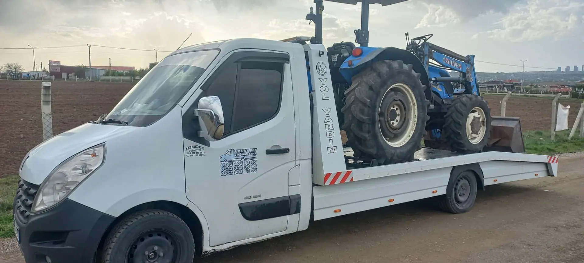 Tarla kenarındaki yolda çekici platformunda taşınan mavi renkli kepçeli traktör