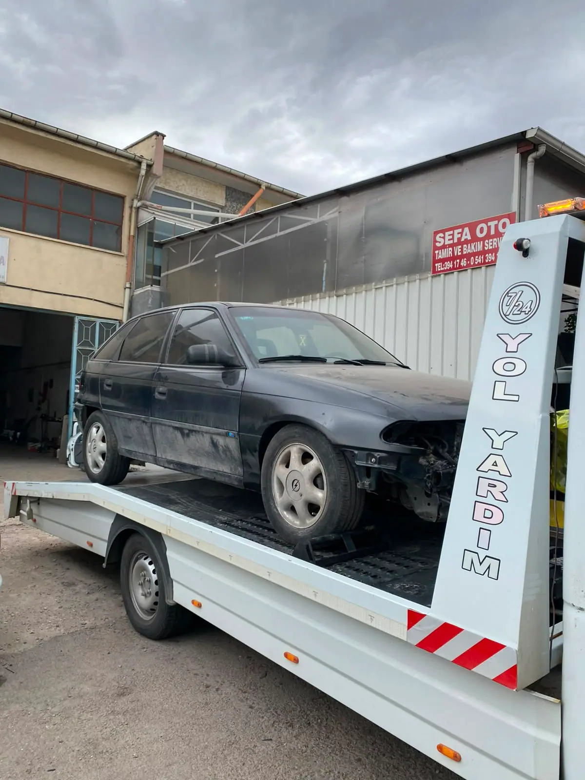 Ön tamponu hasarlı siyah otomobilin oto servis önünde çekici platformunda taşınması