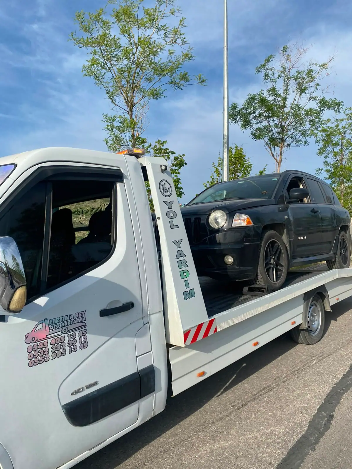 Mavi gökyüzü altında çekici platformunda duran siyah SUV, çekicinin kapısında iletişim bilgileri